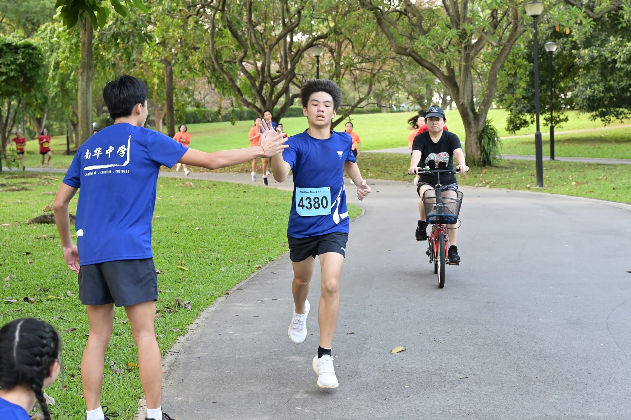 Runner wearing bib number 4380 high-fiving a bystander during a park race, with a cyclist following behind.